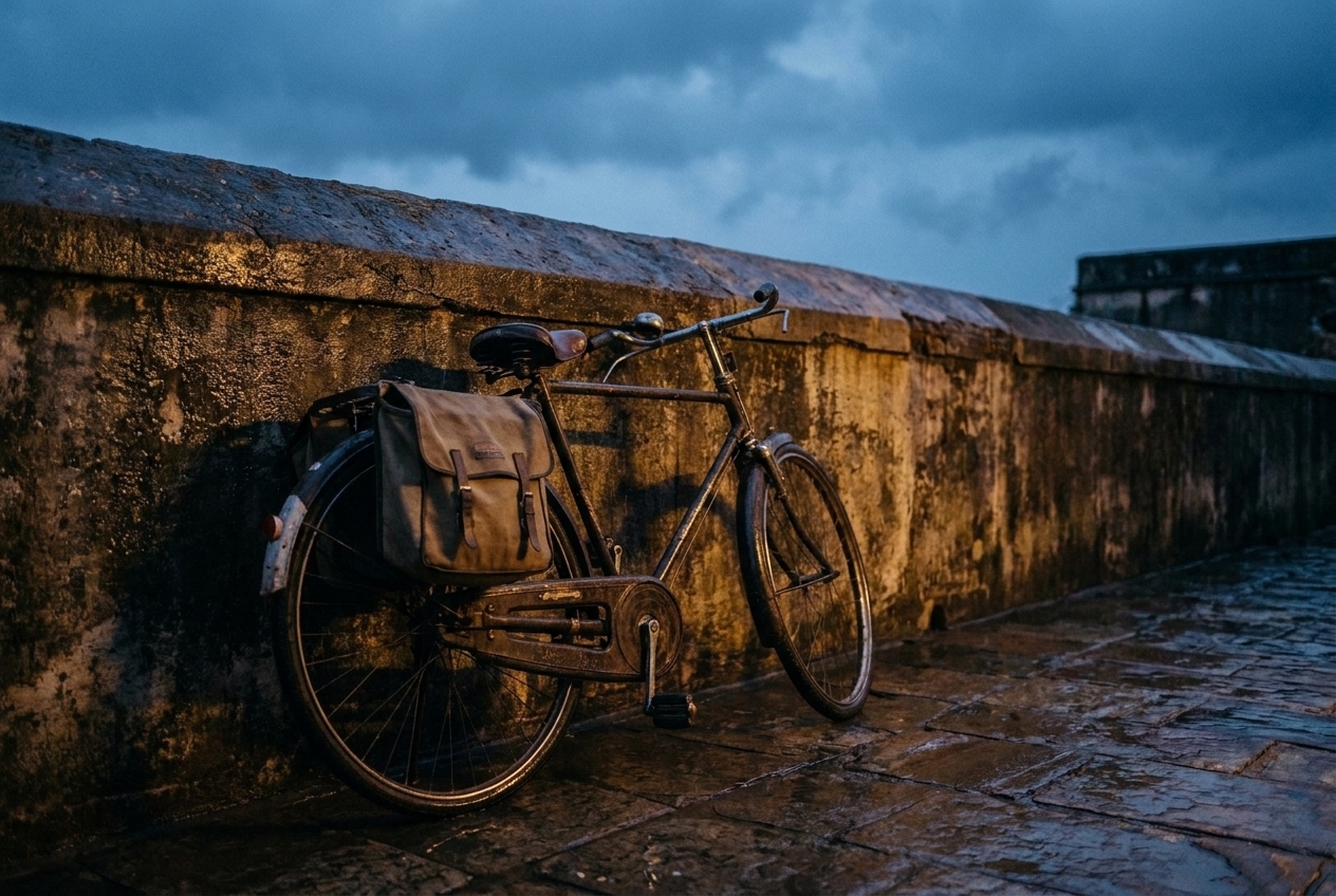 Vintage bicycle with canvas pannier bag parked against a textured stone wall on wet cobblestones at dusk.