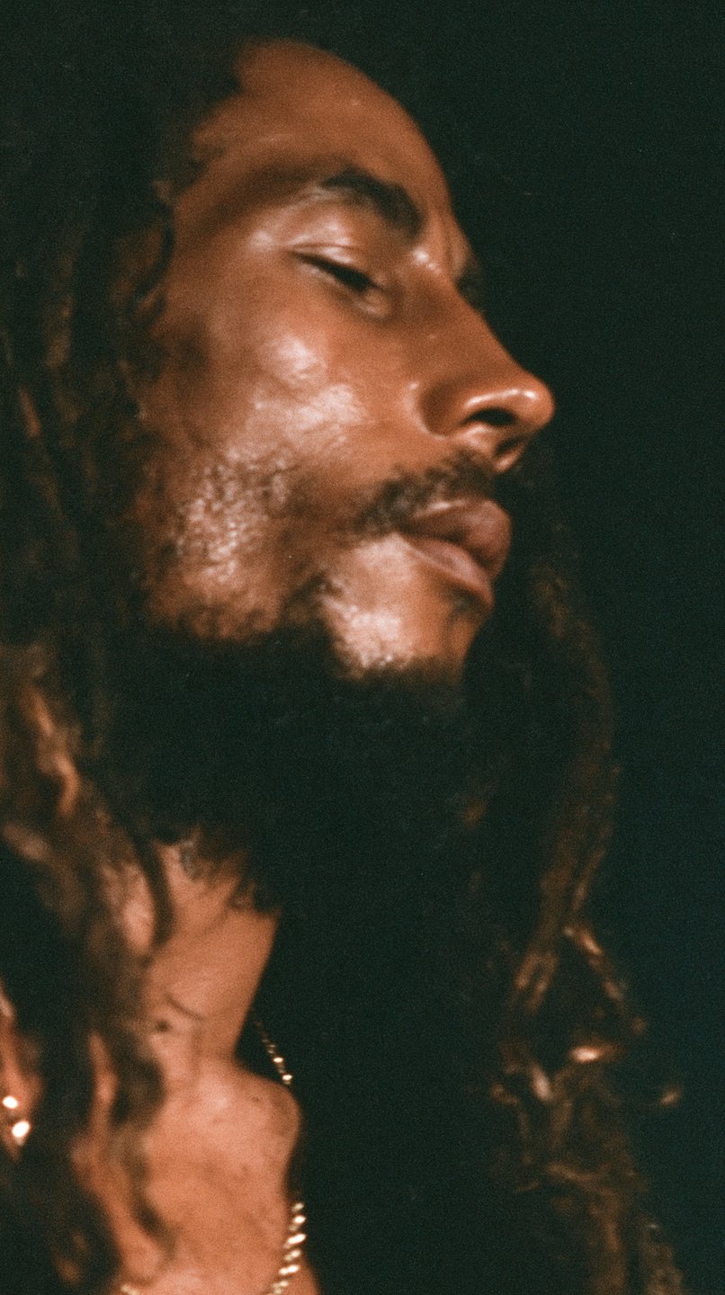 Dreadlocked man in profile with eyes closed, glistening skin, and warm low-key lighting.