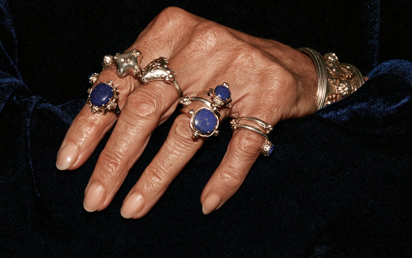 An elegant mature hand adorned with multiple ornate silver and lapis lazuli rings rests on blue velvet.