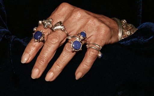 An elegant mature hand adorned with multiple ornate silver and lapis lazuli rings rests on blue velvet.