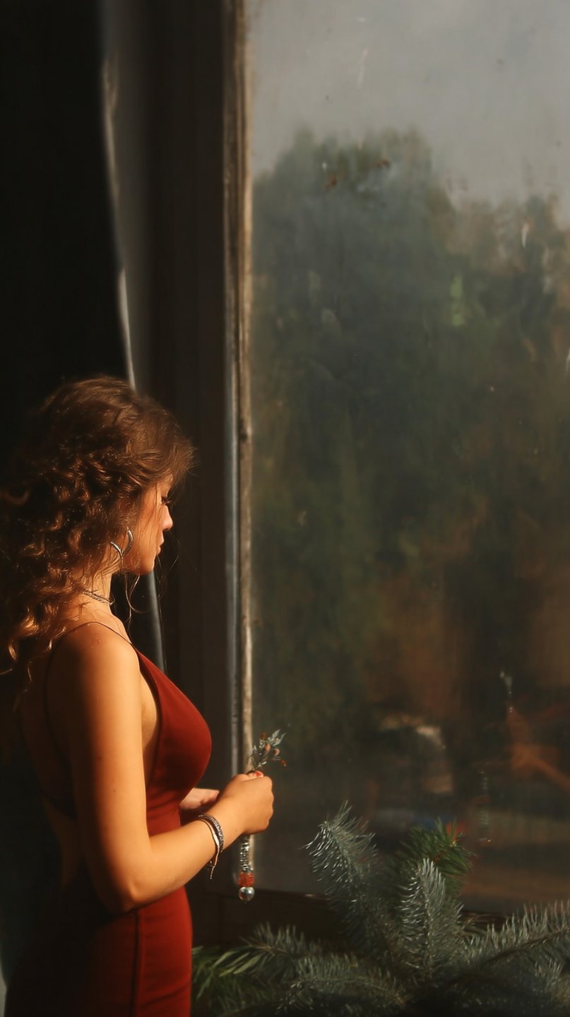 Young woman with curly hair looking out a weathered window while wearing a deep red dress.