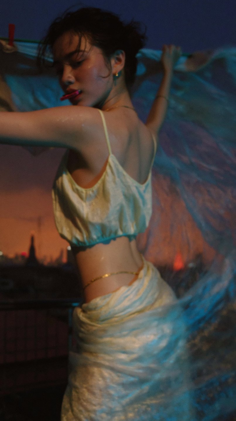 A young woman on a rooftop at dusk holds up a translucent blue sheet against a city skyline.