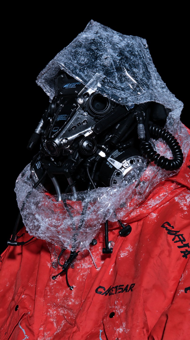 A figure in a red technical jacket wears a black tactical mask under a frosted translucent hood.