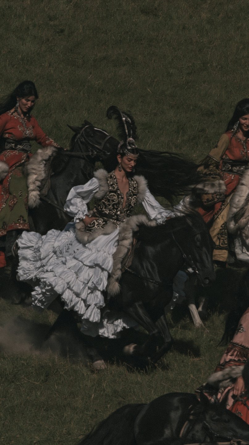 Nomadic horsewomen in ornate traditional attire ride galloping horses across a steep green grassy hillside.