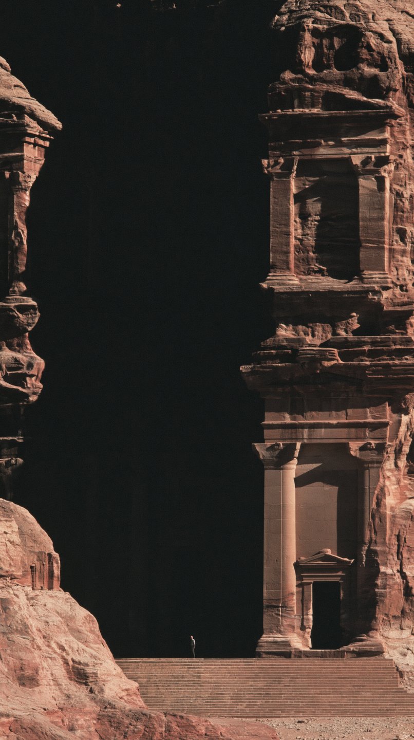 A tiny solitary figure stands on the steps of a monumental sandstone temple carved into a dark canyon.