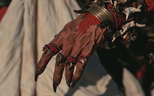 Close-up of a veined hand with long black claws, wearing bone bracelets and beaded rings with red pigment.