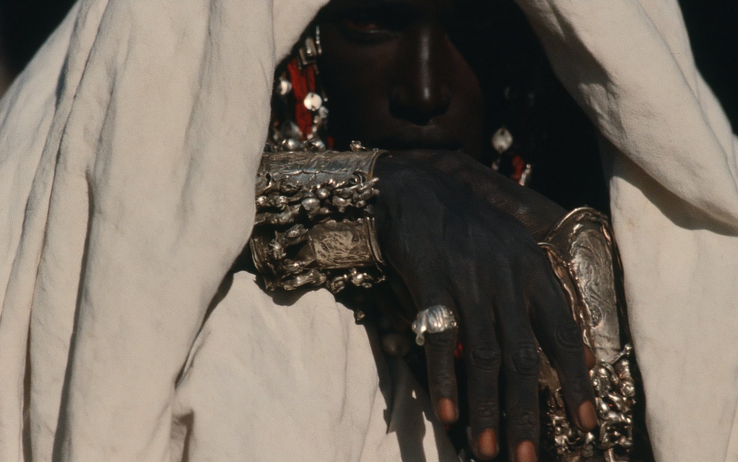 Close-up of a veiled person wearing intricate silver cuffs and rings, their face partially shadowed.