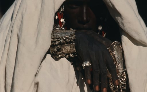 Close-up of a veiled person wearing intricate silver cuffs and rings, their face partially shadowed.