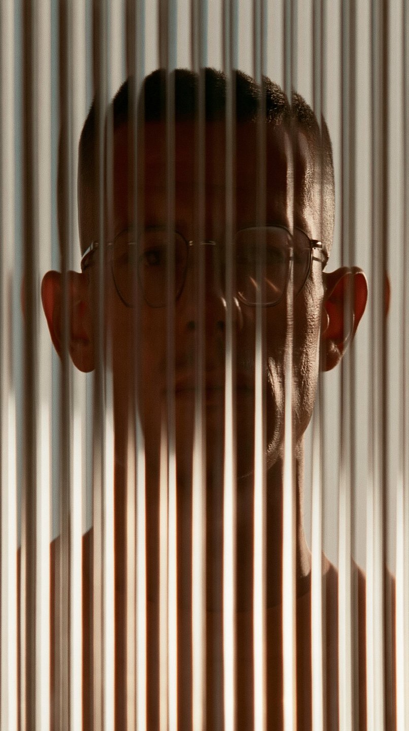 Man with glasses viewed through vertical fluted glass, illuminated by warm rim lighting in deep shadow.