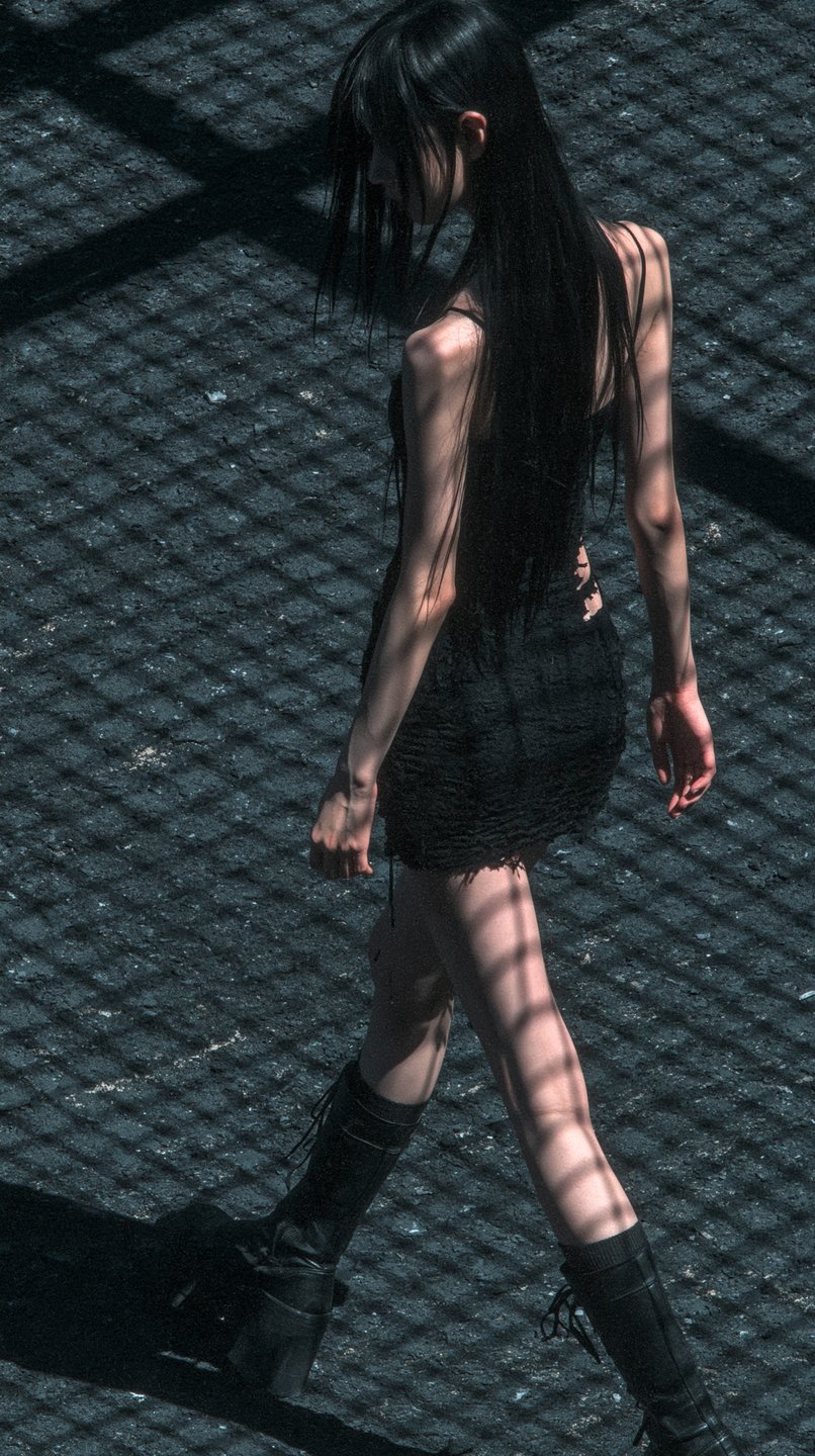 A slender young woman with long black hair walks across a ground covered in grid-like shadows.