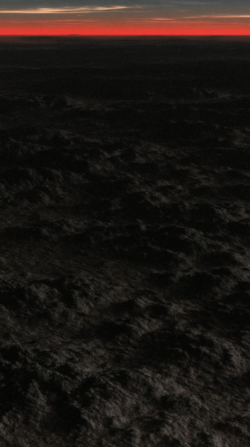 Dark textured volcanic terrain stretches toward a thin, glowing crimson horizon under a black sky.