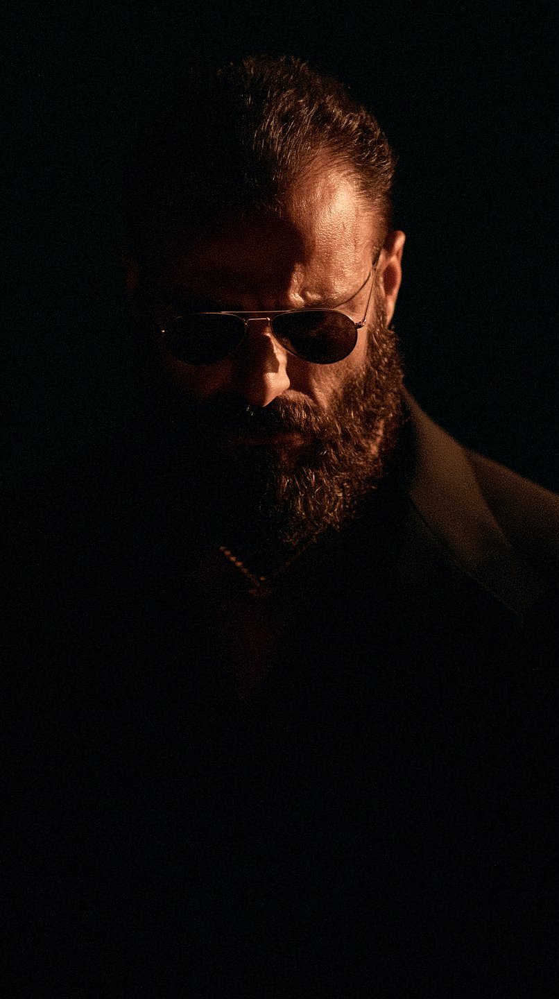 Dramatic low-key portrait of a bearded man wearing aviator sunglasses, illuminated by a single overhead light source.