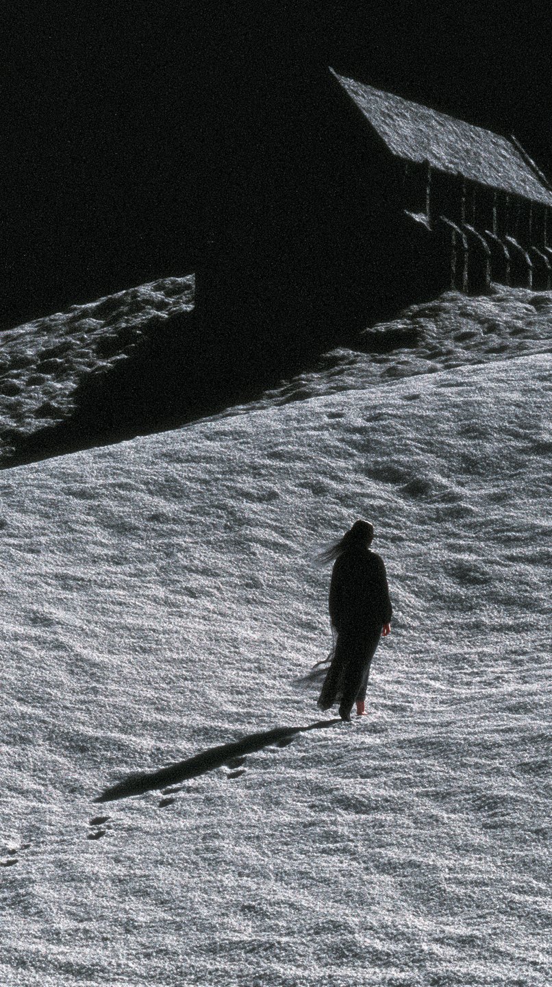 A cloaked woman walks barefoot up a snowy hill toward a dark wooden cabin under moonlight.