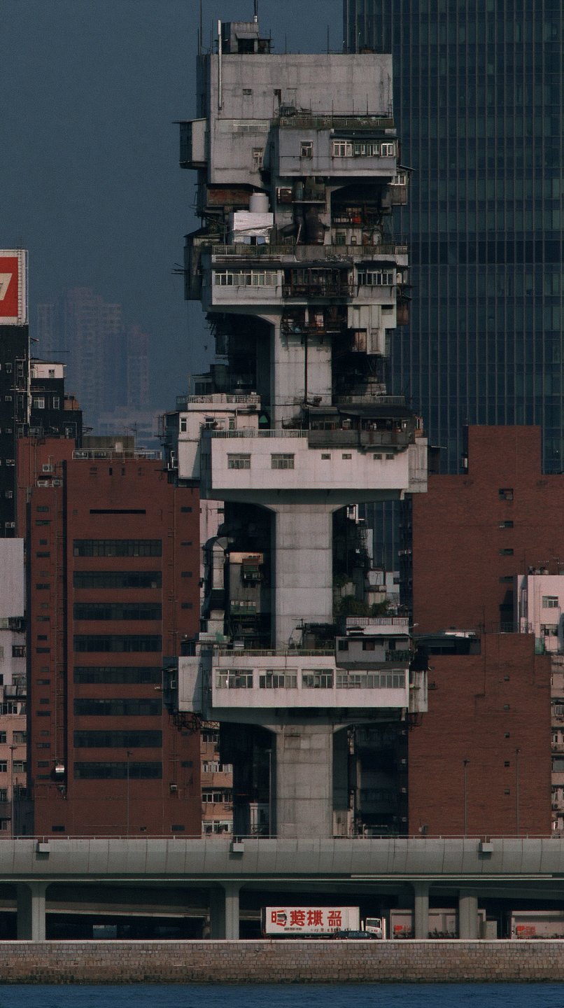 A towering, weathered concrete brutalist building stands vertically amidst modern red and glass skyscrapers in Hong Kong.
