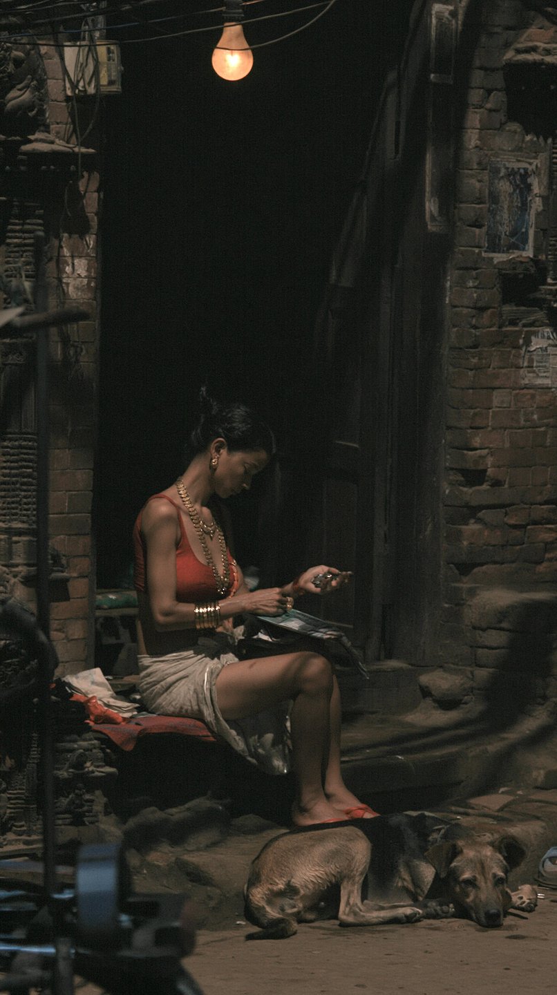 A woman in traditional red attire and gold jewelry sits under a lightbulb in a brick courtyard.