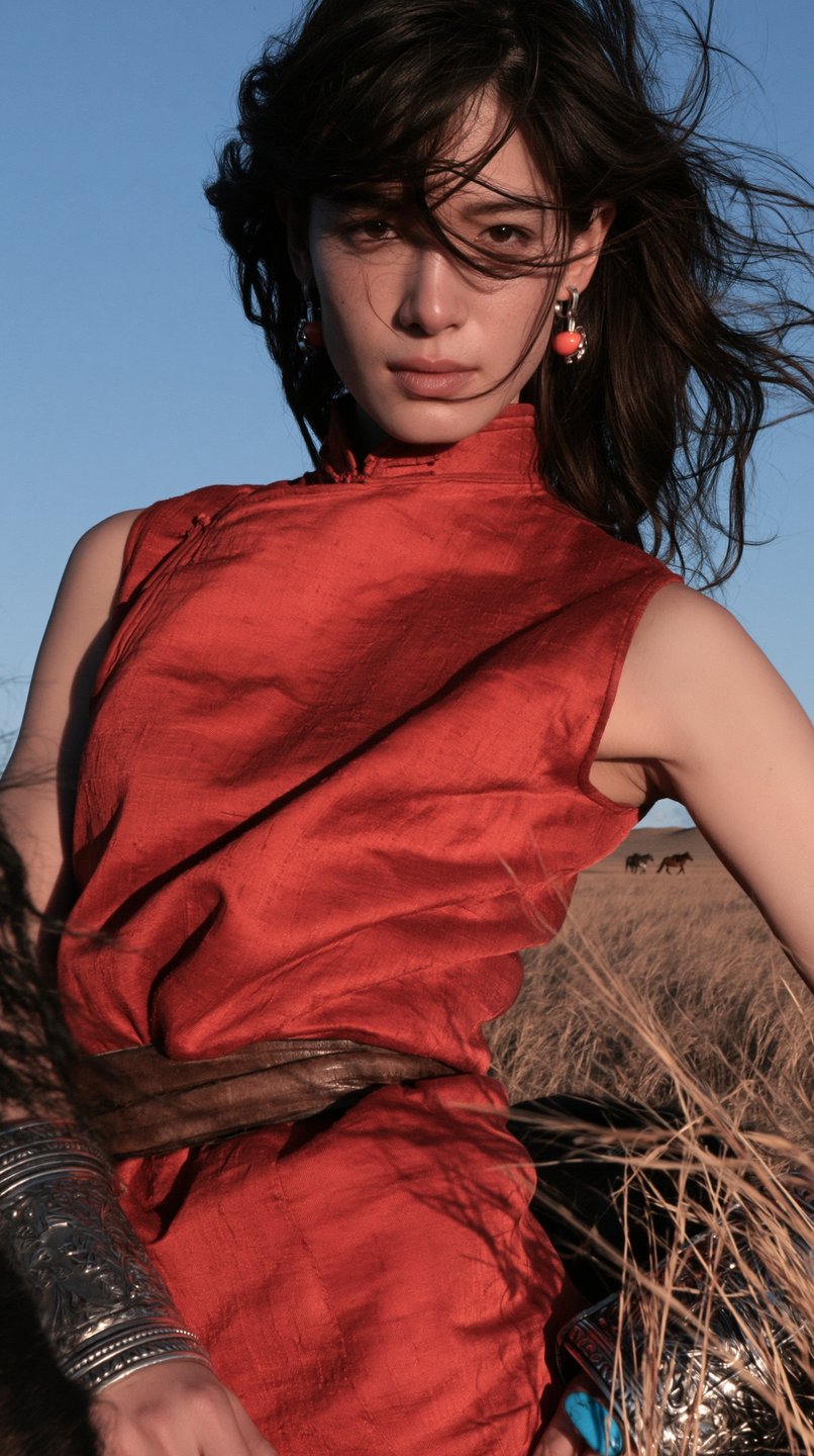 A young woman in a red silk tunic and silver cuffs stands on a windy steppe.