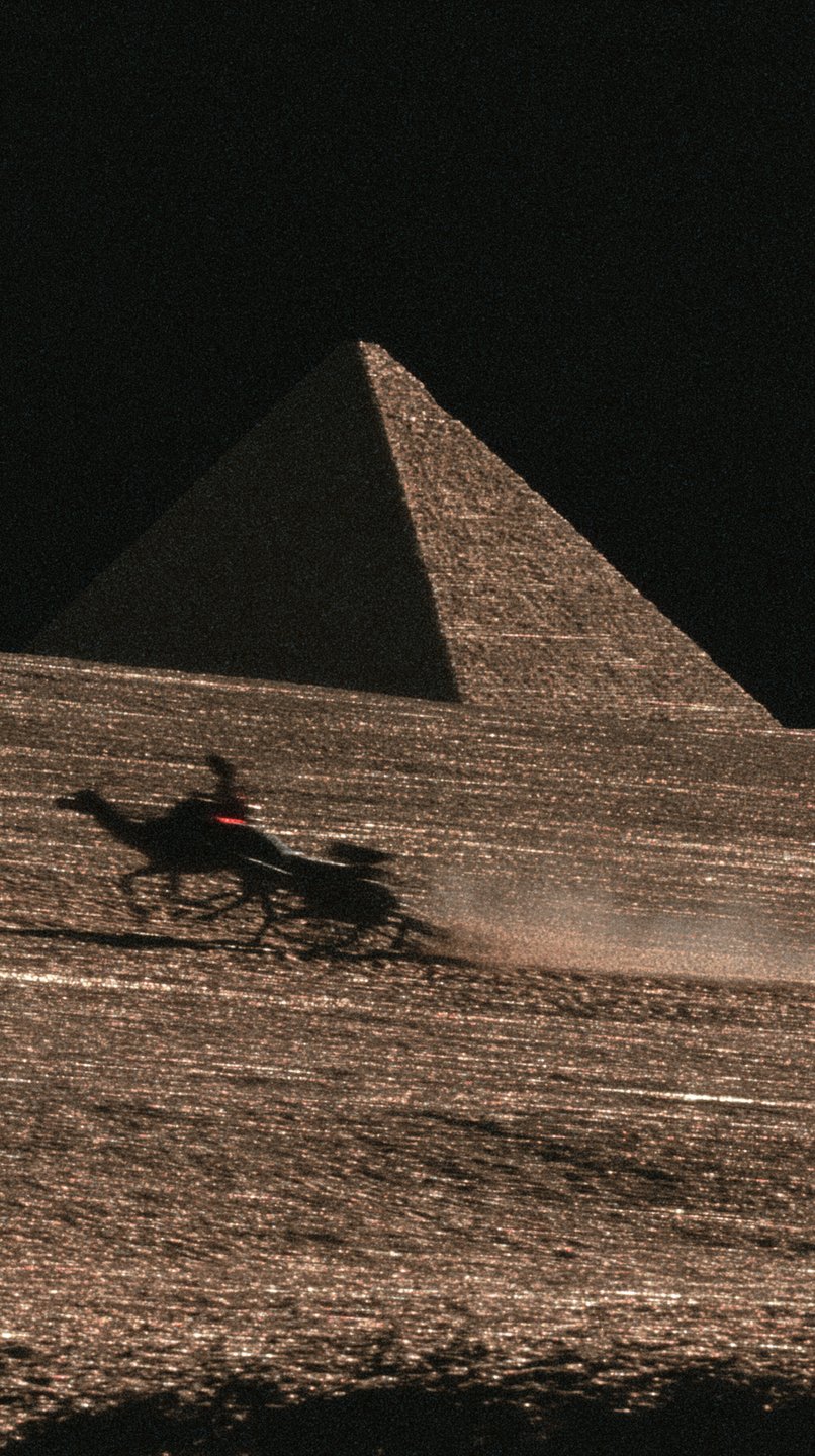 Silhouetted camel rider kicks up dust traversing golden desert sands before a massive pyramid at night.