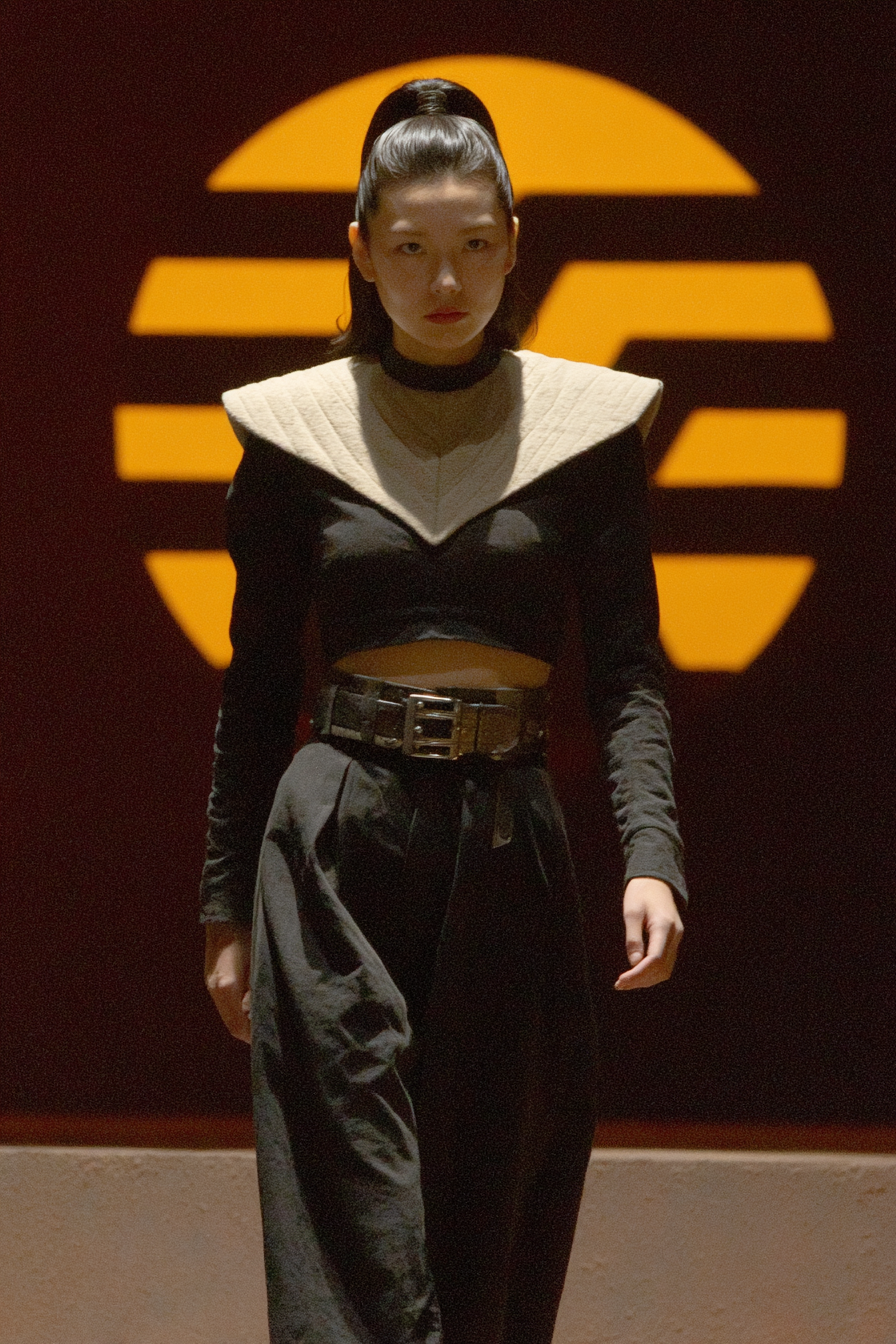 Woman in futuristic black and cream attire stands before a large glowing orange sun-like logo.