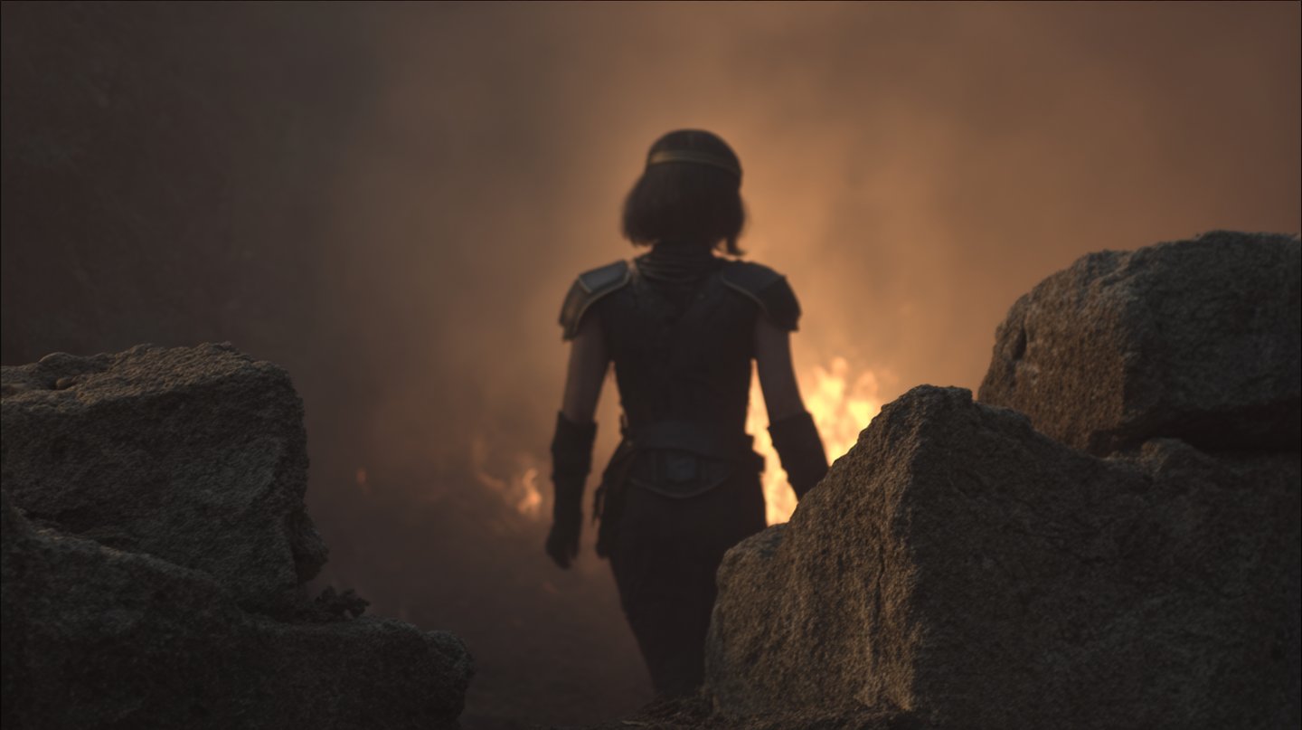 Back view of a female warrior in dark armor walking toward an intense orange fire through rocks.